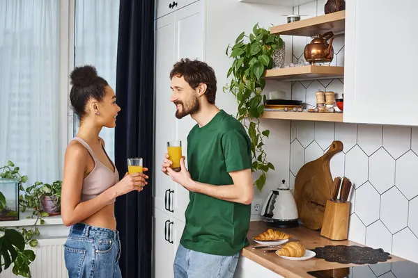 Beautiful joyous diverse couple in homewear drinking fresh orange juice and smiling happily — Stock Photo