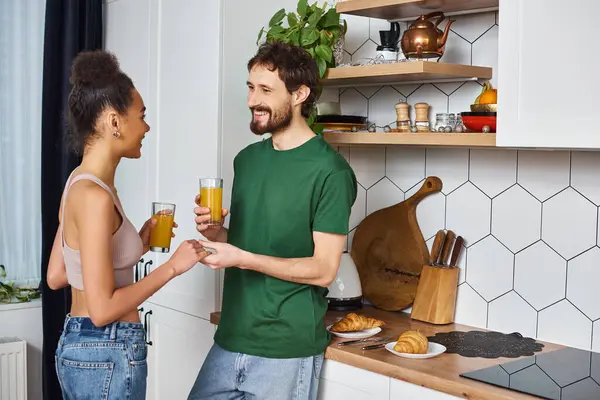 Attraente allegra coppia diversificata in casa bere succo d'arancia e sorridente a vicenda — Foto stock