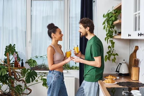 Good looking cheerful interracial couple in casual outfits holding glasses of orange juice at home — Stock Photo