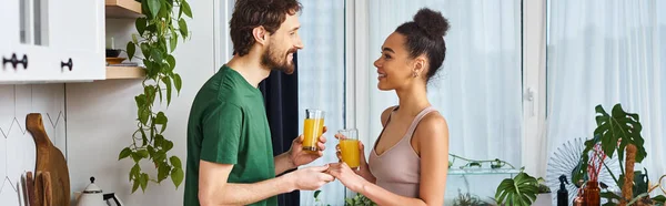 Bella coppia interrazziale allegra che tiene bicchieri di succo d'arancia e sorride felicemente, striscione — Foto stock