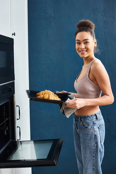 Alegre atractivo africano americana mujer en acogedor hogar hornear croissants y mirando cámara - foto de stock