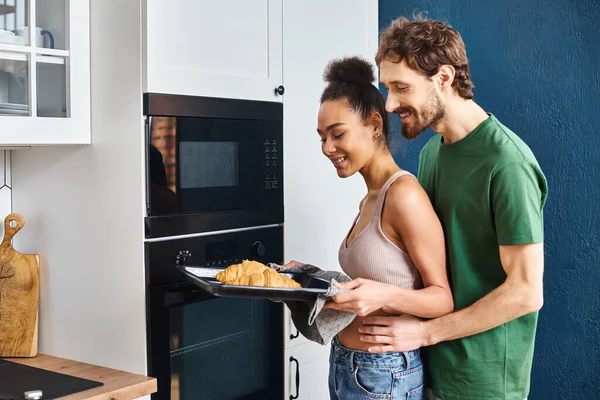 Joyful multicultural couple in homewear using oven to bake delicious croissants for breakfast — Stock Photo