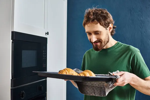 Guapo hombre alegre en ropa de casa usando horno para hornear deliciosos croissants mientras que en la cocina en casa - foto de stock