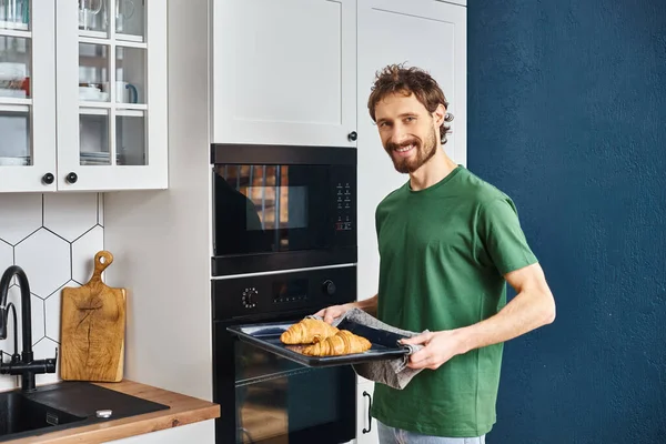 Hombre alegre bien parecido en traje casual usando horno para hornear croissants y sonriendo a la cámara - foto de stock