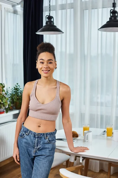 Alluring african american joyous woman in cozy homewear posing next to table and smiling at camera — Stock Photo