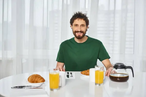 Joyeux homme attrayant en tenue décontractée assis à la table pendant le petit déjeuner et souriant à la caméra — Photo de stock
