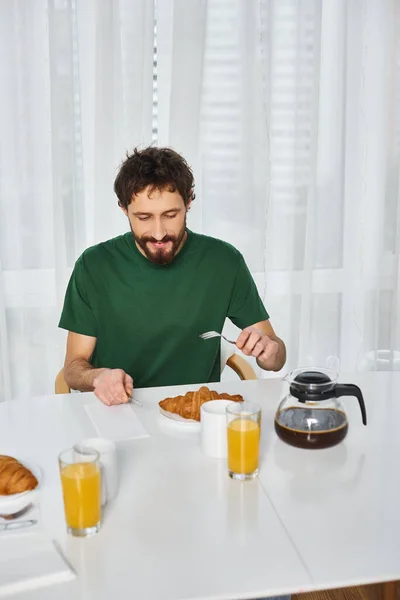 Hombre alegre bien parecido en ropa de casa acogedora disfrutando de delicioso desayuno y sonriendo felizmente en casa - foto de stock