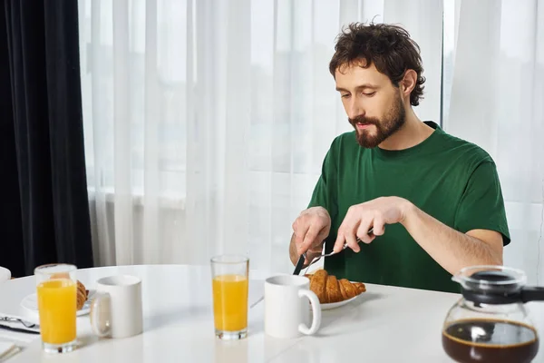 Hombre guapo en camiseta verde casual corte delicioso croissant con cuchillo durante el desayuno en casa - foto de stock