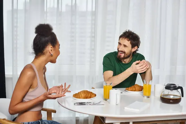 Attractive diverse jolly couple in comfy homewear eating breakfast and smiling at each other — Stock Photo