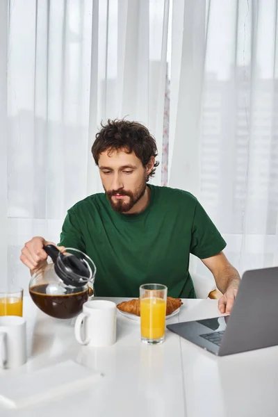 Hombre guapo en ropa cómoda verter un poco de café durante el desayuno con el ordenador portátil en la mesa - foto de stock