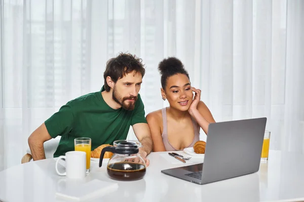 Attractive jolly multicultural couple in cozy homewear watching movies on laptop during breakfast — Stock Photo