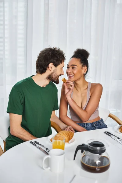 Cheerful beautiful african american woman feeding delicious croissant to her loving boyfriend — Stock Photo