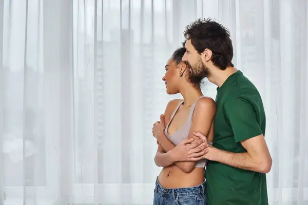 Good looking jolly diverse couple in casual attires hugging warmly while spending time at home — Stock Photo