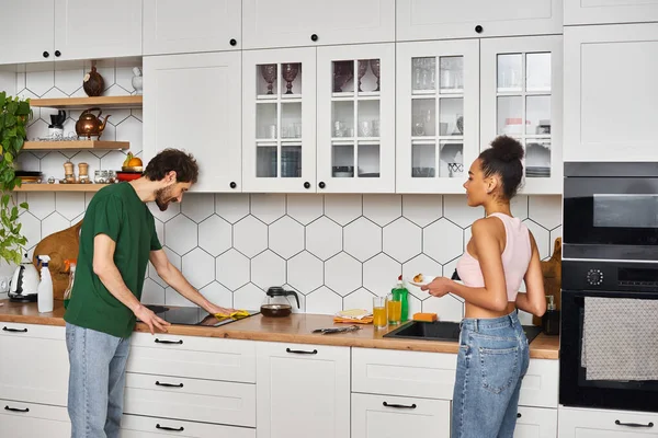 Atractiva pareja interracial alegre en ropa de casa haciendo tareas y limpieza de la cocina después del desayuno - foto de stock