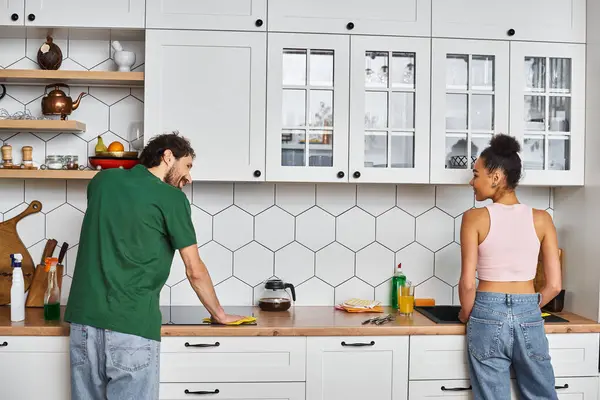 Atractiva pareja multicultural alegre en trajes acogedores limpieza cocina después del desayuno juntos - foto de stock