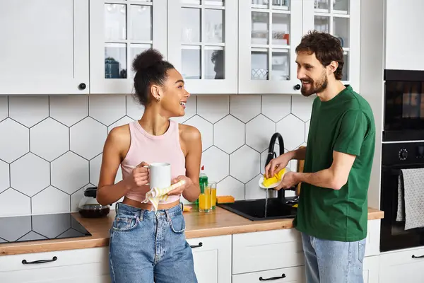 Joyous multicultural beautiful couple in homewear washing dishes and smiling at each other at home — Stock Photo