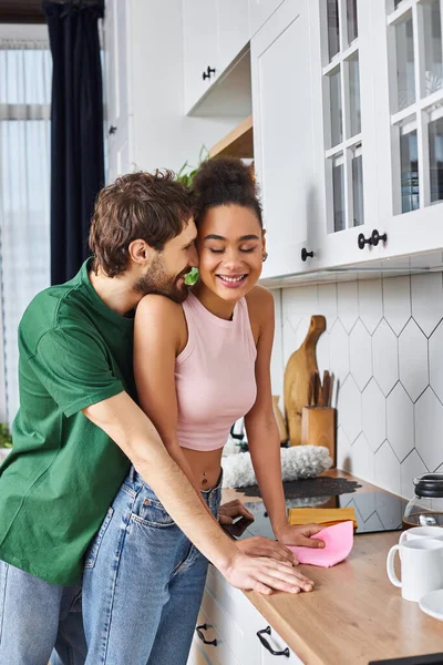 Atractiva pareja multicultural alegre en la ropa de casa abrazándose amorosamente durante la limpieza de primavera en casa - foto de stock