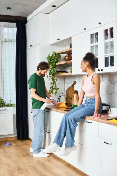 Appealing jolly african american woman sitting on counter and watching her boyfriend doing chores — Stock Photo
