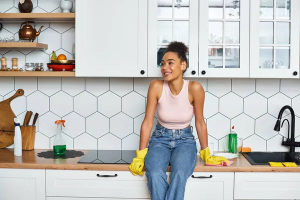 Alegre atractiva mujer afroamericana en ropa de casa sentado en el mostrador de la cocina y mirando hacia otro lado - foto de stock