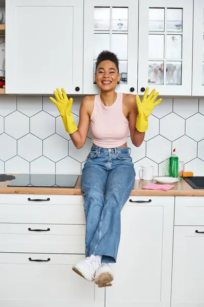 Alluring jolly african american woman in homewear sitting on kitchen counter and smiling at camera — Stock Photo