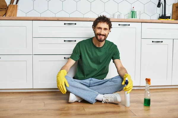 Handsome cheerful man in dark green t shirt sitting on floor and smiling at camera, spring cleaning — Stock Photo