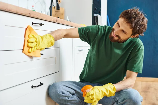Hombre guapo alegre en guantes usando trapo y limpiador para lavar el mostrador de la cocina, limpieza de primavera - foto de stock