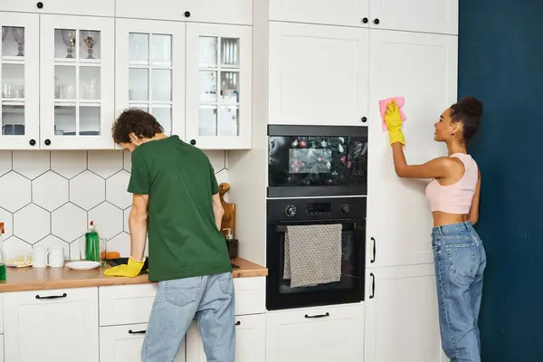 Весёлая многорасовая пара в повседневных нарядах, выполняющая свою работу на кухне во время весенней уборки. — стоковое фото