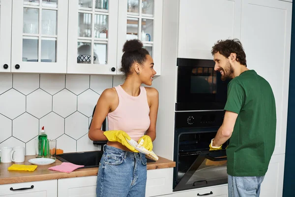 Alegre pareja multirracial de buen aspecto en atuendos cotidianos limpieza de su cocina en casa - foto de stock