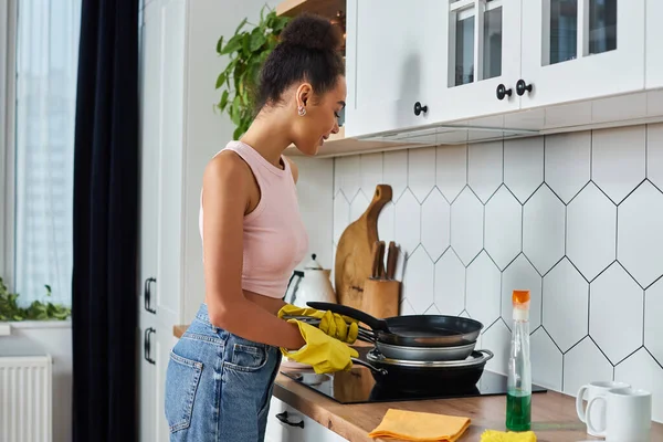Привлекательная африканская женщина в кастрюлях для чистки одежды и других принадлежностях на кухне — стоковое фото