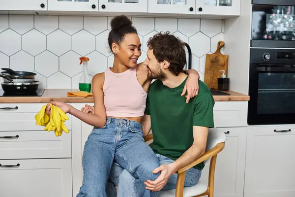Loving cheerful multiracial couple sitting on chair and hugging with rags and gloves in hands — Stock Photo