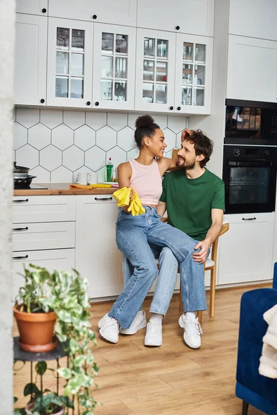 Joyful beautiful diverse couple in homewear sitting on chair with rags and gloves, spring cleaning — Stock Photo