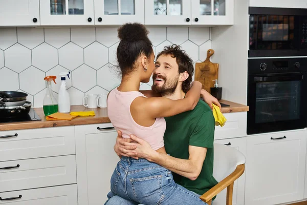 Alegre casal multicultural em homewear sentado na cadeira e abraçando de forma fascinante, limpeza de primavera — Fotografia de Stock