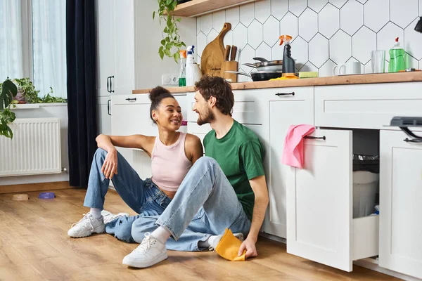 Appealing multiracial joyous couple in homewear sitting on floor and smiling at each other — Stock Photo