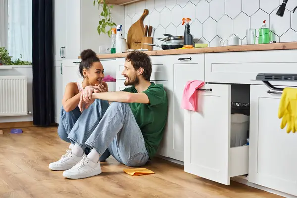 Attractive cheerful diverse couple in cozy homewear sitting on floor and smiling at each other — Stock Photo