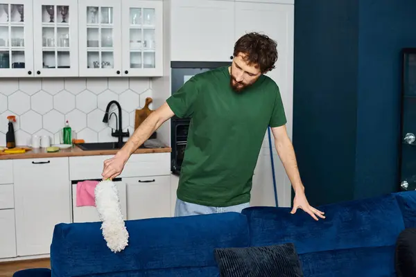 Hombre guapo en cómodo traje casual limpieza sofá con plumero con cocina de fondo - foto de stock
