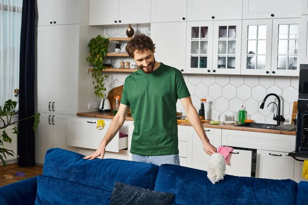 Allegro bell'uomo in accogliente divano pulizia casalinga con spolverino e sorridente felicemente mentre a casa — Foto stock