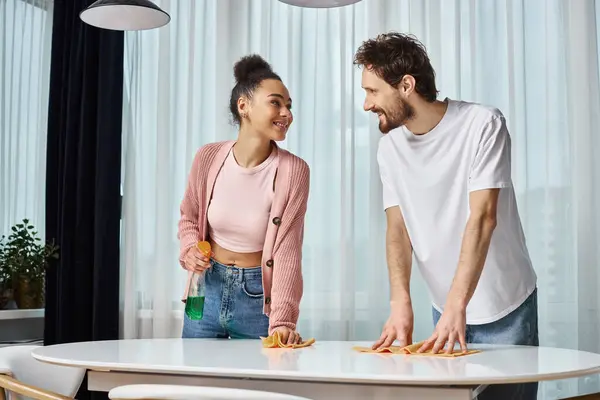 Allegro attraente coppia diversificata in comodi casalinghi sorridenti a vicenda durante la pulizia a casa — Foto stock