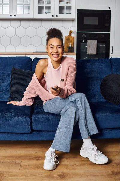 Beautiful joyful african american woman in casual attire sitting on sofa and watching TV at home — Stock Photo