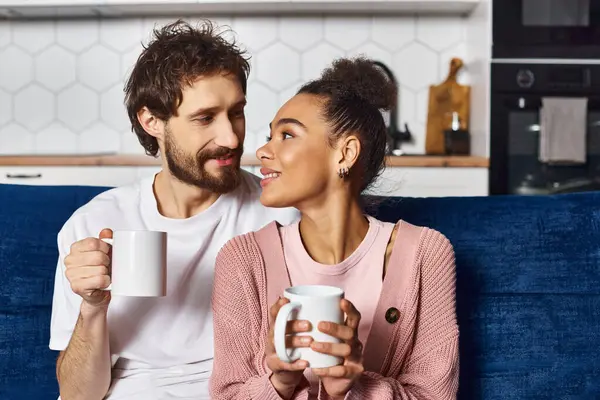 Good looking jolly diverse couple in casual attire sitting on sofa and drinking coffee at home — Stock Photo