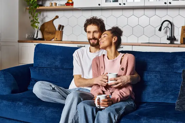 Joyous interracial couple in homewear drinking coffee together while sitting on sofa at home — Stock Photo