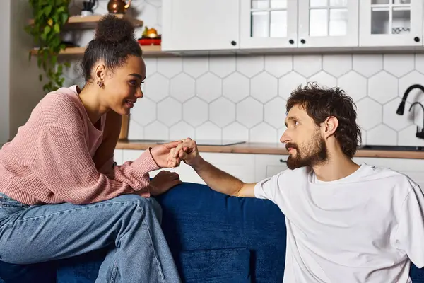 Atractiva pareja multirracial alegre en acogedora ropa de casa sentado en el sofá y cogido de la mano en casa — Stock Photo