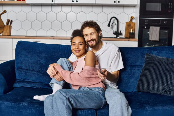 Joyful multicultural couple in comfy homewear hugging on sofa and looking at camera at home — Stock Photo
