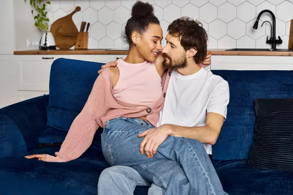 Cheerful multiracial couple in comfy homewear sitting on sofa together and smiling at each other — Stock Photo