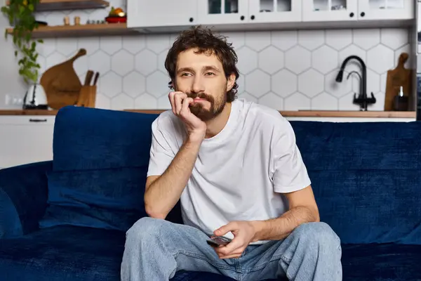 Bored handsome man in cozy homewear sitting on sofa with remote control in hand and watching TV — Stock Photo