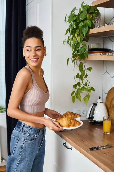 Joyeuse femme afro-américaine en tenue décontractée tenant des assiettes avec des croissants et détournant les yeux — Photo de stock