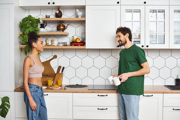 Boa aparência casal diversificado em homewear acolhedor olhando uns para os outros, enquanto na cozinha, os amantes em casa — Fotografia de Stock