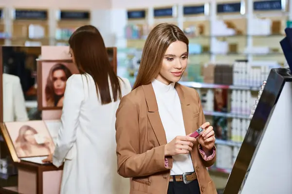 Atractivo cliente rubio elegir lápiz labial con su amigo borroso en el fondo en la tienda de cosméticos - foto de stock