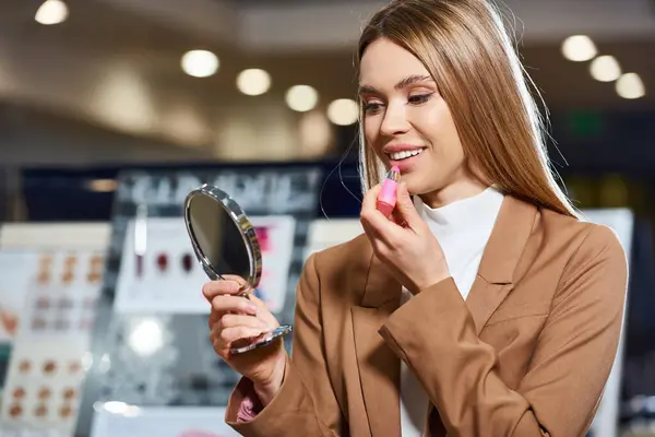 Appealing jolly woman in business casual attire choosing new lipstick while in cosmetics store — Stock Photo