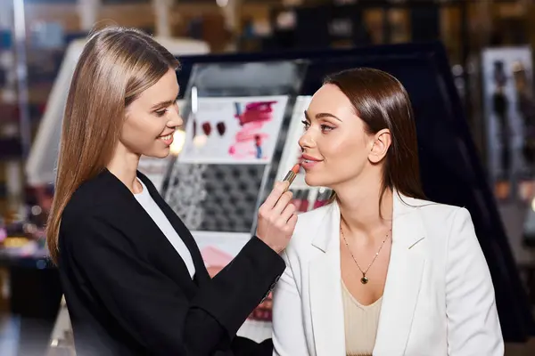 Beautiful shop assistant in black blazer applying lipstick on her customer while in cosmetics store — Stock Photo