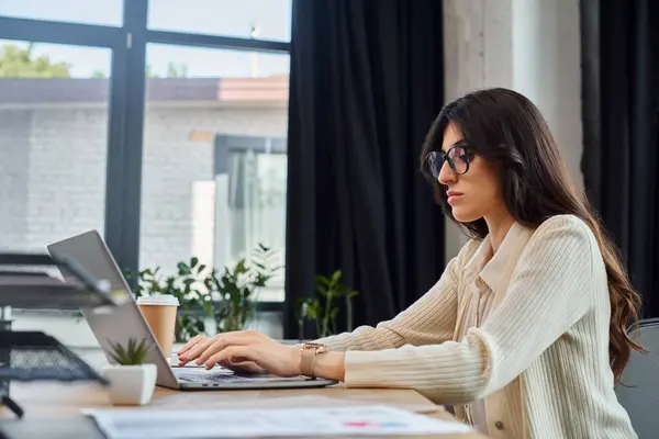 Une femme d'affaires est assise à un bureau, concentrée sur son écran d'ordinateur portable, dans un cadre de bureau moderne avec des articles liés à la franchise. — Photo de stock